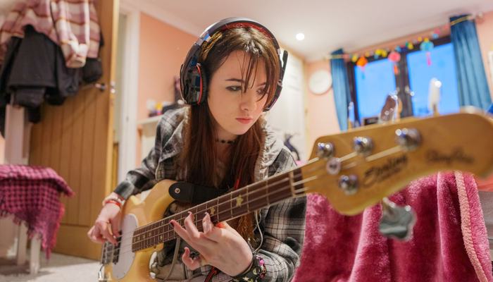 Young Carer Meghan, sitting on her bedroom floor, wearing headphones and playing her bass guitar.