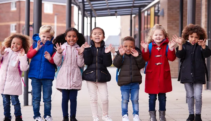 Seven young children standing in a row outdoors under a covered walkway, all raising their hands with fingers spread. Children wear colourful winter clothing, with a brick building and windows visible in the background.