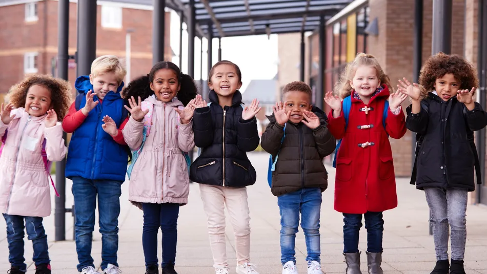 Seven young children standing in a row outdoors under a covered walkway, all raising their hands with fingers spread. Children wear colourful winter clothing, with a brick building and windows visible in the background.