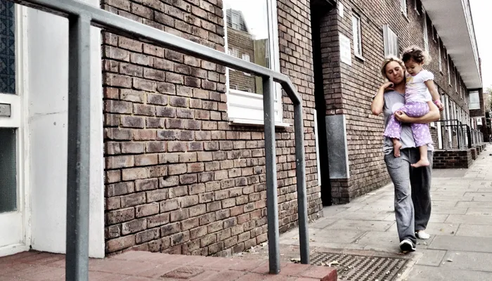 Person carrying a child while walking along next to a brick building with multiple windows and doors and metal railings. The scene highlights an urban environment, possibly local authority accommodation.