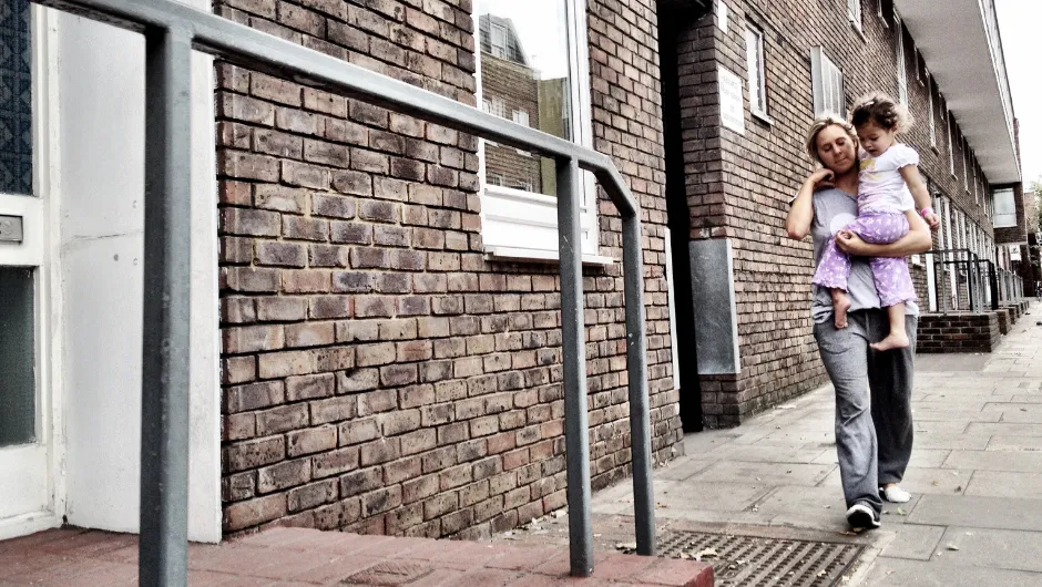 Person carrying a child while walking along next to a brick building with multiple windows and doors and metal railings. The scene highlights an urban environment, possibly local authority accommodation.