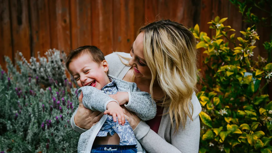 A woman holding a young child outdoors in a garden setting with wooden fence and green foliage in background. Both are smiling, highlighting a tender moment between them.
