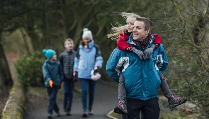 Two adults and three children walking outdoors on a paved path surrounded by trees and greenery. A person in a blue jacket carries a child on their back in the foreground, while three others, dressed in winter clothing, walk behind them.