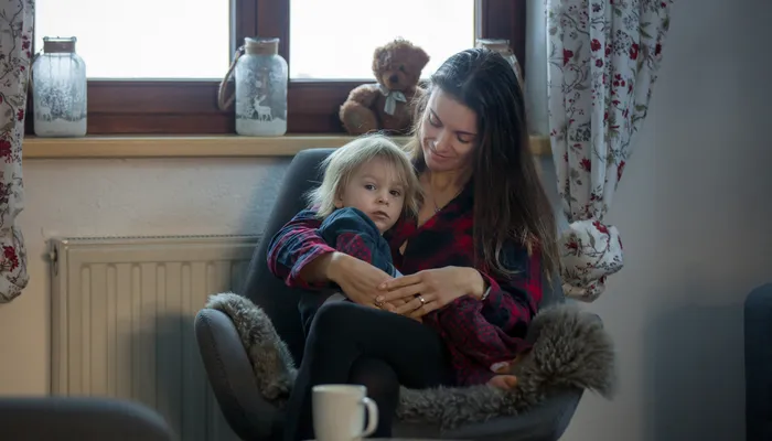 A woman and child sitting together in a cozy armchair near a window with floral curtains, creating a warm, intimate setting. The scene includes decorative jars and a teddy bear on the windowsill, with a small round table and a white mug in the foreground.