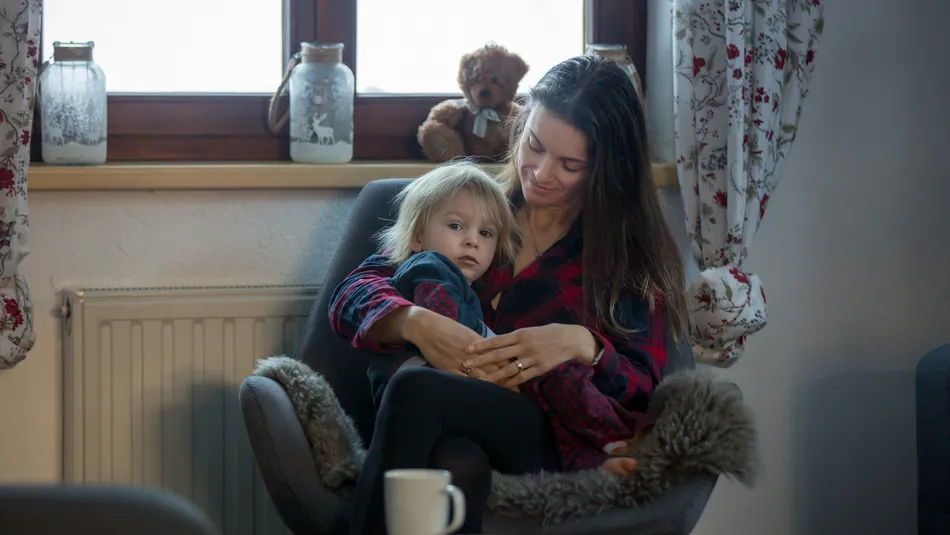 A woman and child sitting together in a cozy armchair near a window with floral curtains, creating a warm, intimate setting. The scene includes decorative jars and a teddy bear on the windowsill, with a small round table and a white mug in the foreground.