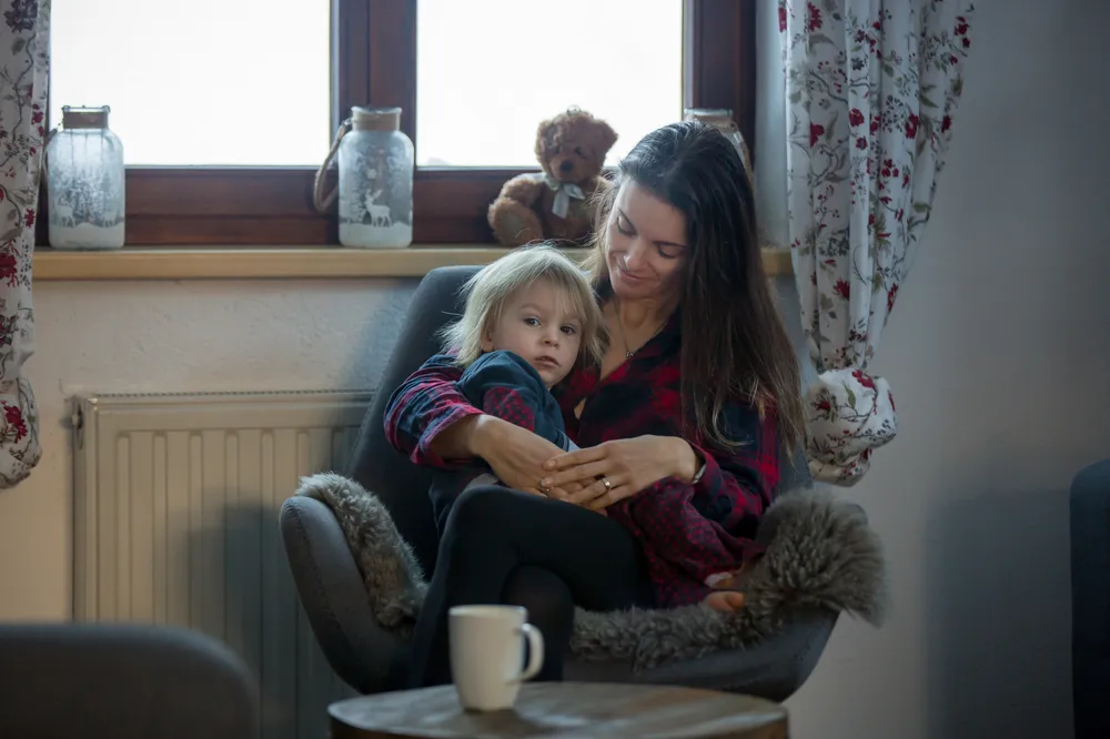 A woman and child sitting together in a cozy armchair near a window with floral curtains, creating a warm, intimate setting. The scene includes decorative jars and a teddy bear on the windowsill, with a small round table and a white mug in the foreground.