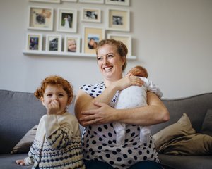 Mother with her two children in the living room of their home. The mother is holding a baby and the other little girl is laughing and looking at the camera while sucking her thumb.