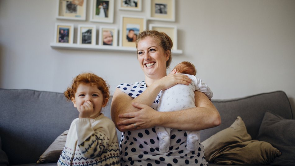 Mother with her two children in the living room of their home. The mother is holding a baby and the other little girl is laughing and looking at the camera while sucking her thumb.