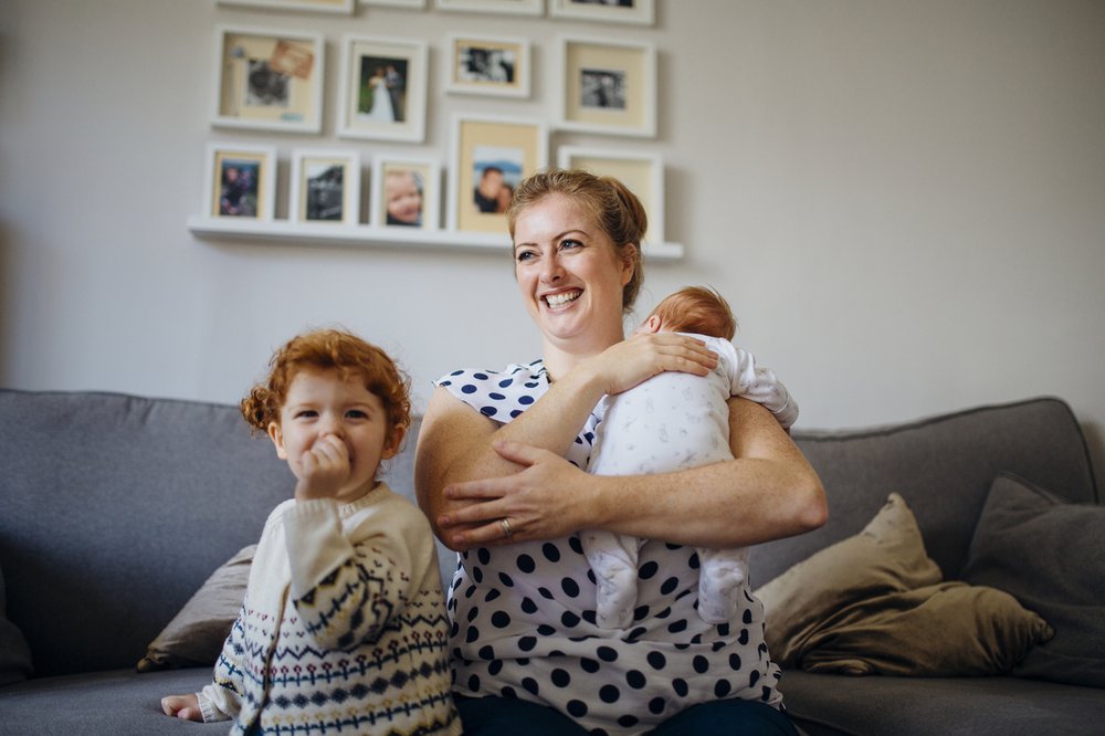 Mother with her two children in the living room of their home. The mother is holding a baby and the other little girl is laughing and looking at the camera while sucking her thumb.