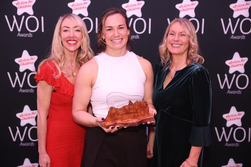 Three women standing in front of a backdrop with the Action for Children logo across it; the central woman is holding an award