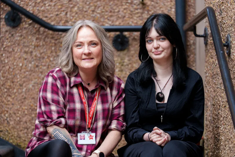 An older woman and younger woman looking at the camera, the older woman has grey hair and a patterned shirt and the younger woman has black hair and black clothing