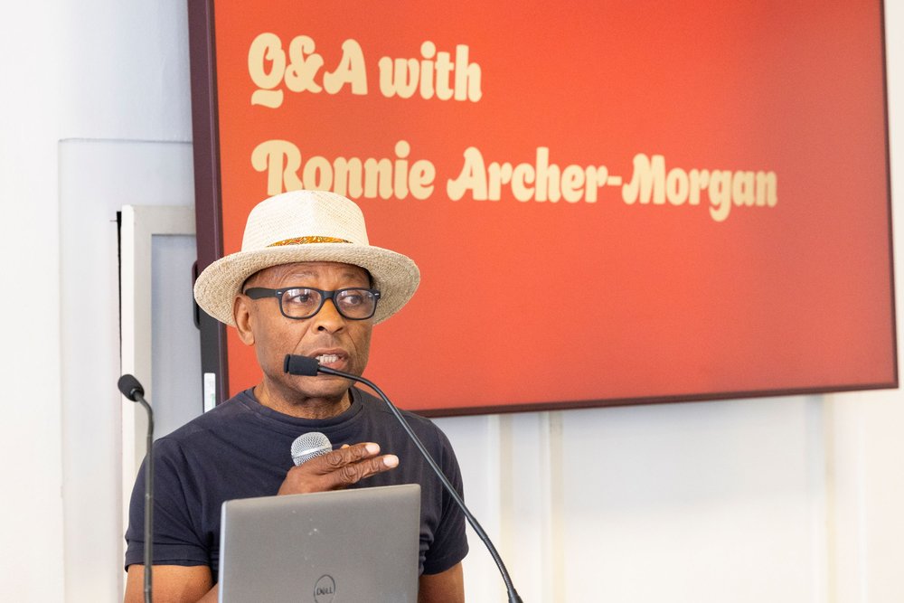 A man speaking at a microphone stand wearing a white hat