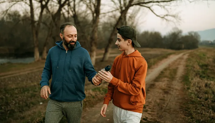 Two people walking along a path surrounded by leafless trees and grass, with one person handing a water bottle to the other. The scene suggests a casual walk or light exercise during a cool season, indicated by their hooded sweatshirts and relaxed posture