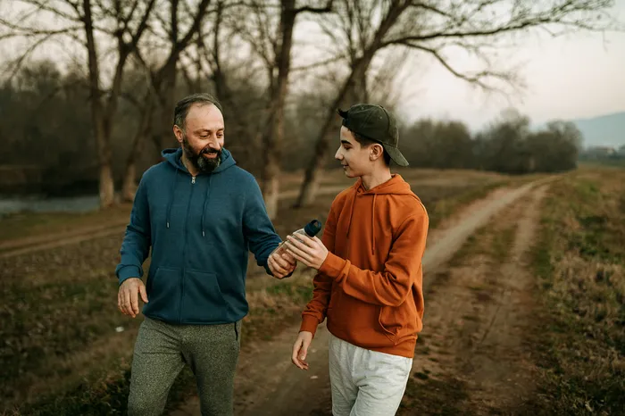 Two people walking along a path surrounded by leafless trees and grass, with one person handing a water bottle to the other. The scene suggests a casual walk or light exercise during a cool season, indicated by their hooded sweatshirts and relaxed posture