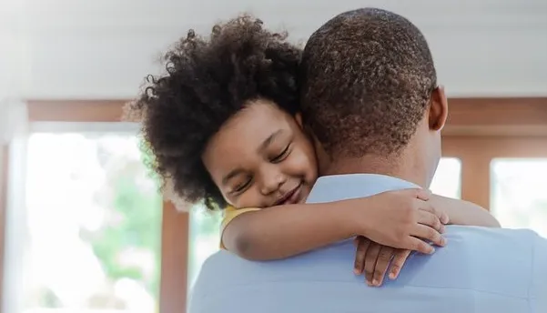 A person with short hair wearing a light blue shirt holding a child with curly hair, who has their arms wrapped around the person's neck. The background includes large windows with natural light, suggesting a warm, indoor setting.