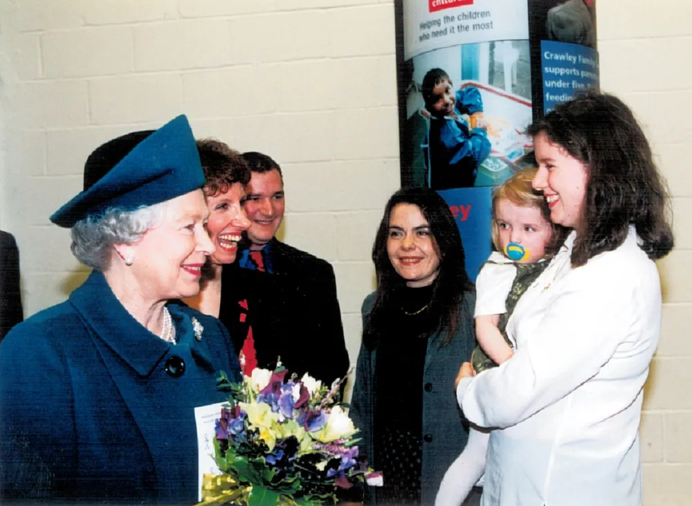 Queen Elizabeth II in a blue outfit and hat holding a bouquet of flowers and a group of people, including a woman holding a child. The background features informational posters, suggesting a public or community event focused on support or charity.