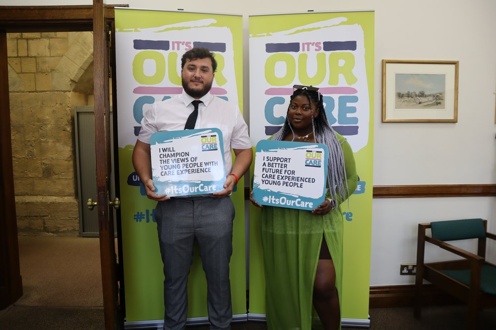 Rachel and Geroge stand holding signs about #ItsOurCare