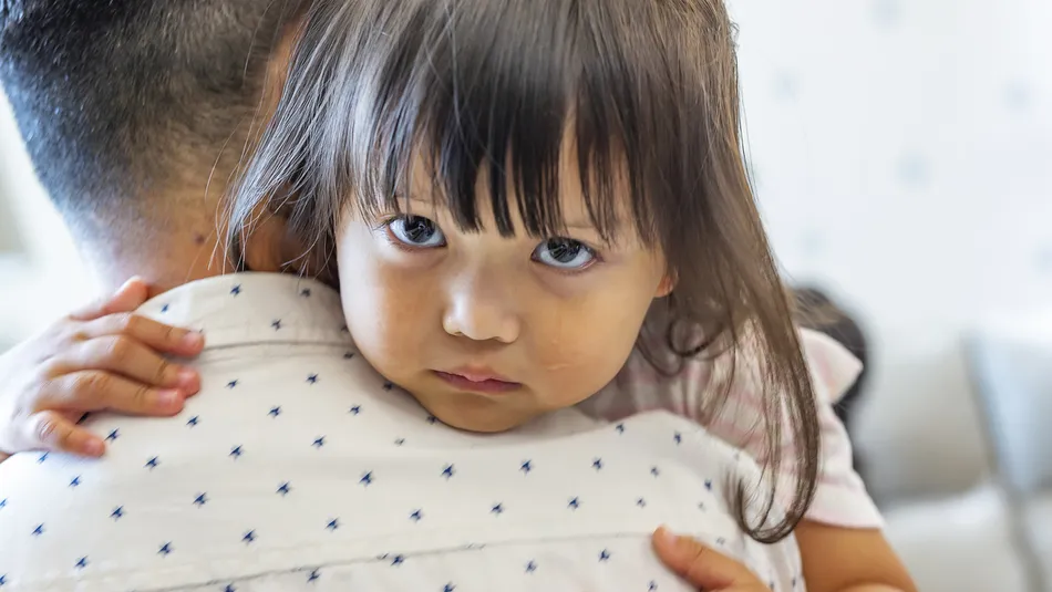 A sad looking child with tears hugging an adult, showing close physical affection and emotional connection. The child’s small hands grip the adult’s back, who is wearing a white shirt with small blue patterns, highlighting a moment of comfort and care.