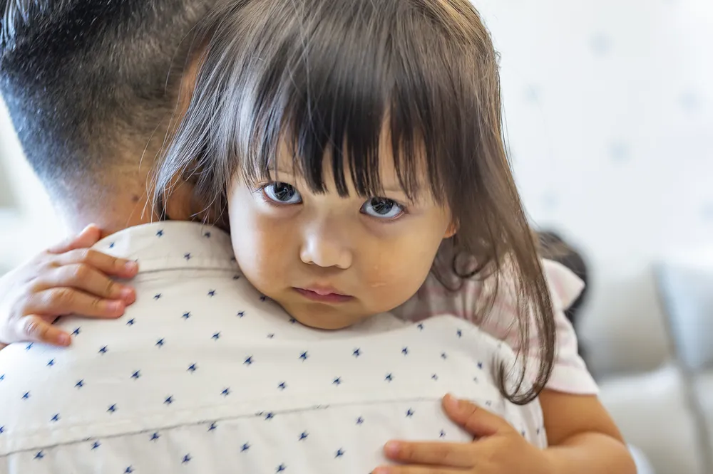 A sad looking child with tears hugging an adult, showing close physical affection and emotional connection. The child’s small hands grip the adult’s back, who is wearing a white shirt with small blue patterns, highlighting a moment of comfort and care.