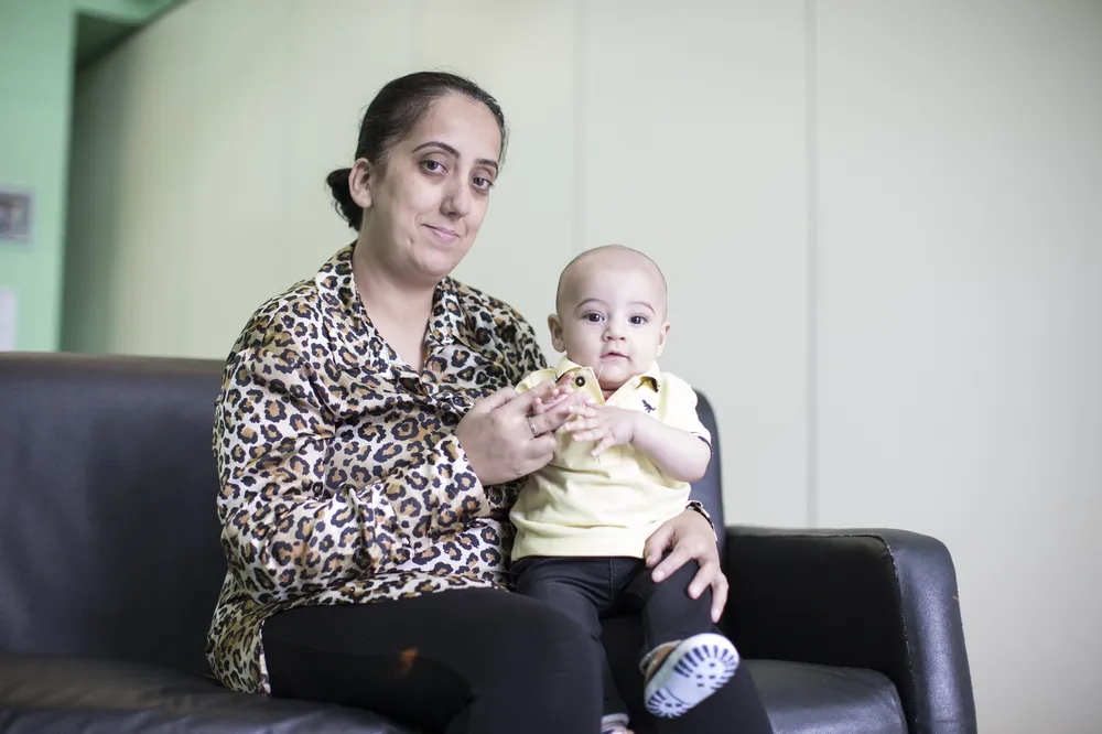 Shadia, a young woman in a anmal print shirt sits on a black sofa holding her baby son who also looking in to the camera