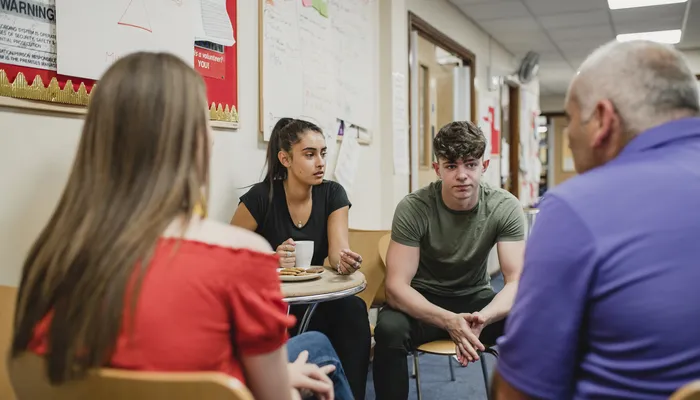 Four people engaged in a discussion while seated in a hallway with bulletin boards and posters on walls. The scene suggests an informal or counselling setting.