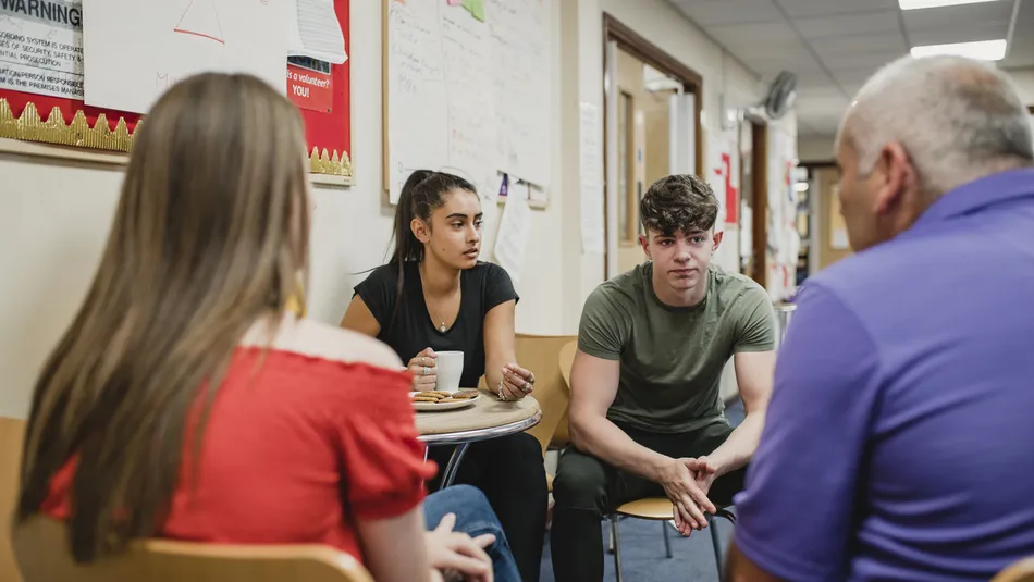 Four people engaged in a discussion while seated in a hallway with bulletin boards and posters on walls. The scene suggests an informal or counselling setting.