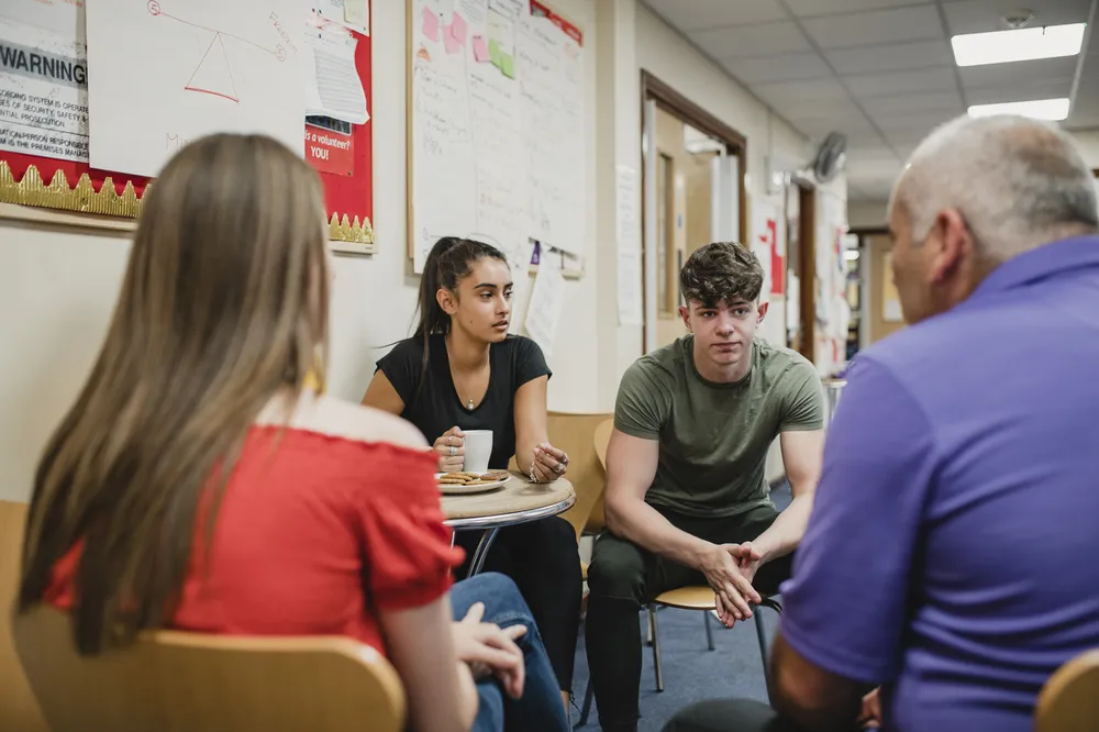 Four people engaged in a discussion while seated in a hallway with bulletin boards and posters on walls. The scene suggests an informal or counselling setting.