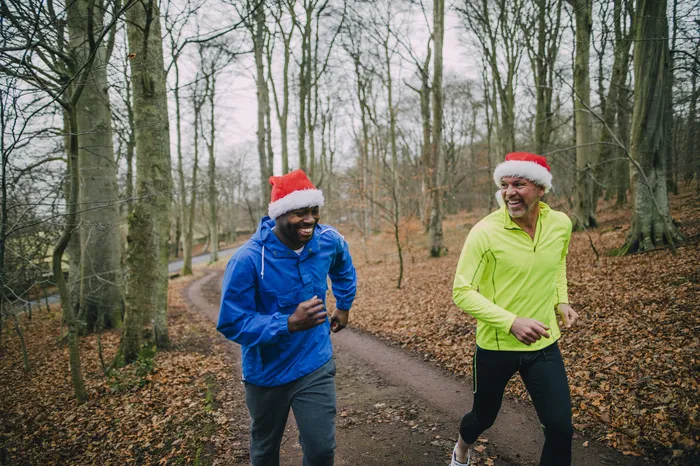 Two people jogging on a forest trail during late autumn, wearing bright athletic jackets—one blue and one neon yellow—and red Santa hats. The scene highlights outdoor exercise in a natural setting with bare trees and fallen leaves covering ground.