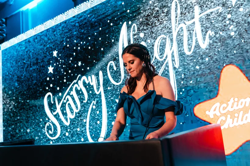 Kat Snoob, behind a DJ booth, wearing headphones and a blue off the shoulder dress