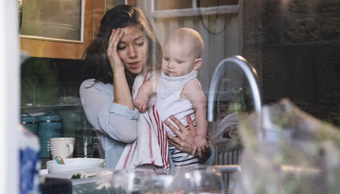 Stressed young mother in kitchen carrying baby on her hip