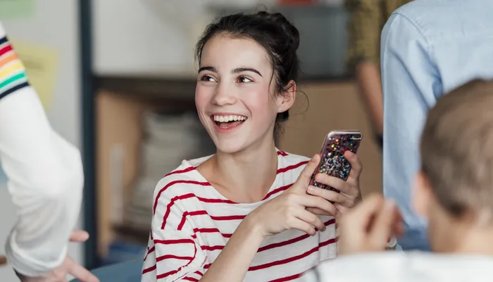 A young person wearing a striped red and white T-shirt  holding a smartphone with a colourful case, engaged in conversation with others. The setting appears casual and social, with blurred individuals in the background suggesting a group interaction.