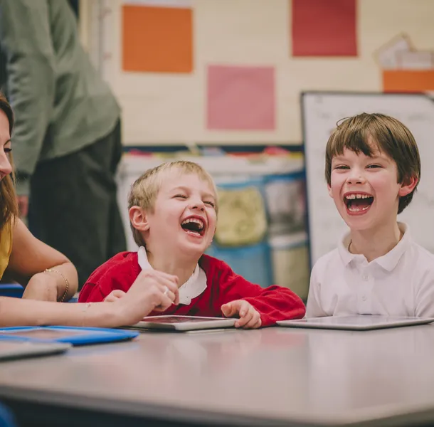 Young children and an adult sitting around a table in a classroom setting, engaged in an activity with tablets. Colourful posters and a whiteboard are visible in the background, indicating an educational environment focused on interactive learning.