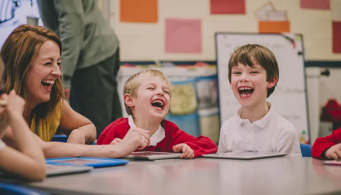 Young children and an adult sitting around a table in a classroom setting, engaged in an activity with tablets. Colourful posters and a whiteboard are visible in the background, indicating an educational environment focused on interactive learning.