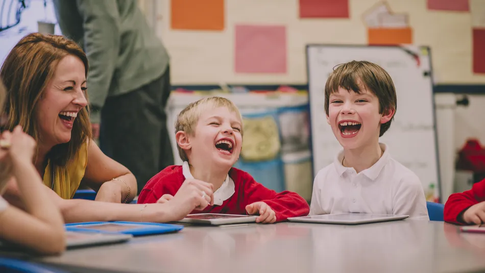 Young children and an adult sitting around a table in a classroom setting, engaged in an activity with tablets. Colourful posters and a whiteboard are visible in the background, indicating an educational environment focused on interactive learning.
