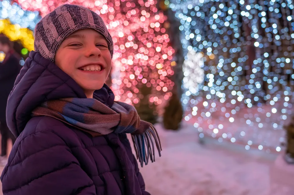 A smiling child dressed in a purple winter coat, multicoloured scarf, and knitted hat standing outdoors at night. Background features vibrant, blurred holiday lights in red, blue, and white, creating a festive atmosphere.
