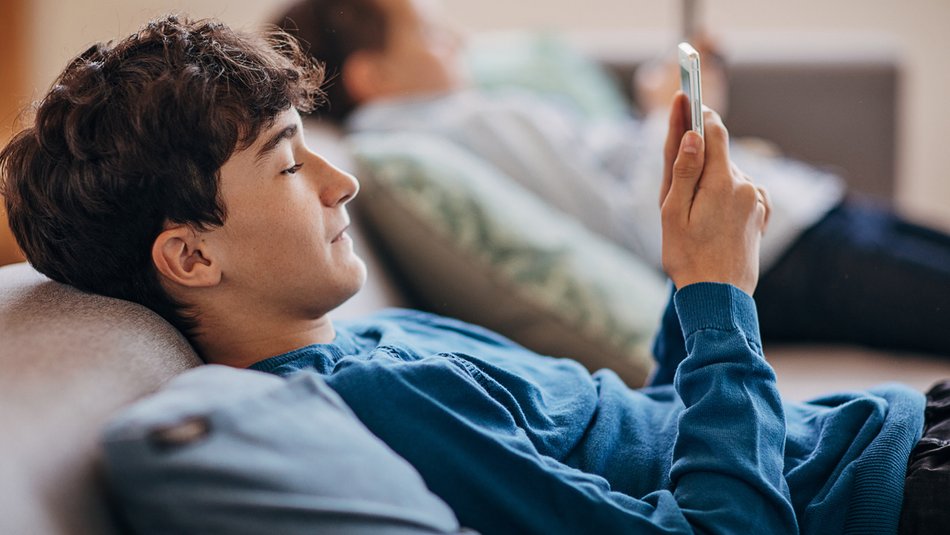 A person in the foreground reclining on a sofa holding and looking at a smartphone. Another person is in the blurred background also holding and looking at a mobile phone. The setting appears relaxed and casual, suggesting leisure or downtime at home.
