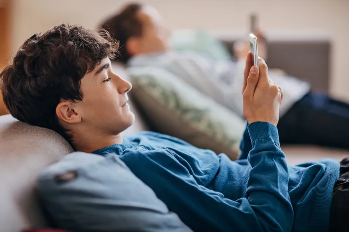 A person in the foreground reclining on a sofa holding and looking at a smartphone. Another person is in the blurred background also holding and looking at a mobile phone. The setting appears relaxed and casual, suggesting leisure or downtime at home.