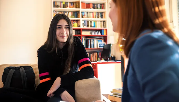 An adult and a teenager engaged in a conversation in a room with bookshelves in the background. One person wears a black sweater with pink and orange stripes on the sleeves, while the other wears a blue jacket, suggesting an informal or counseling setting