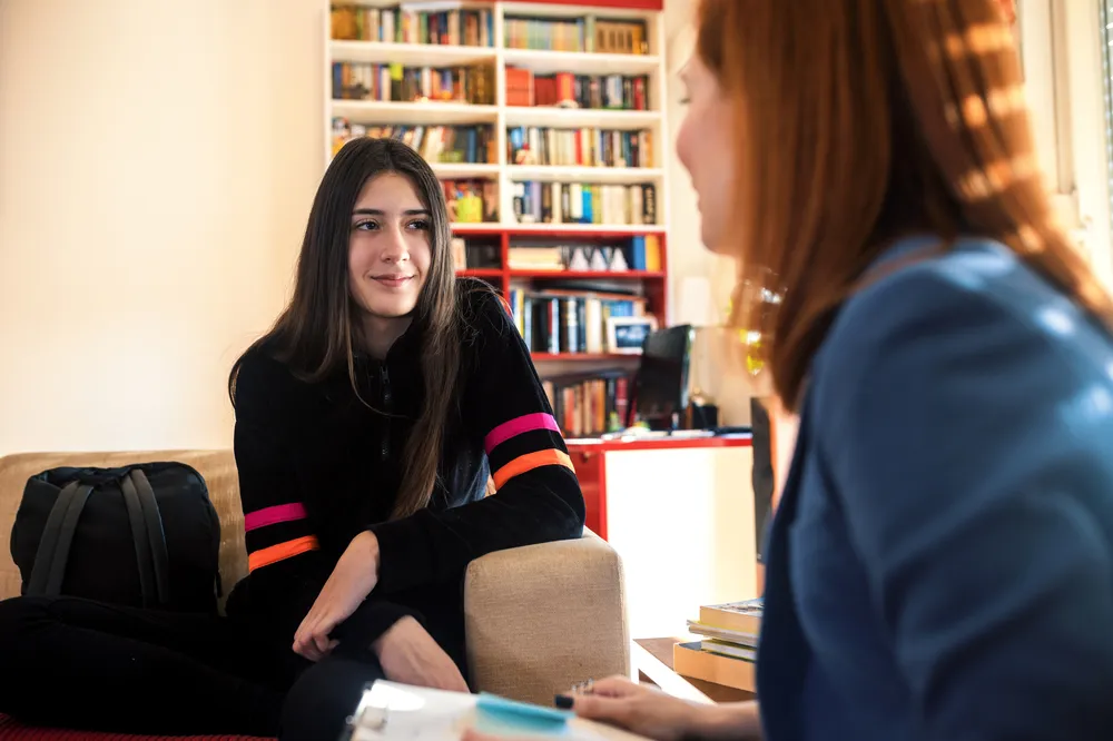 An adult and a teenager engaged in a conversation in a room with bookshelves in the background. One person wears a black sweater with pink and orange stripes on the sleeves, while the other wears a blue jacket, suggesting an informal or counseling setting