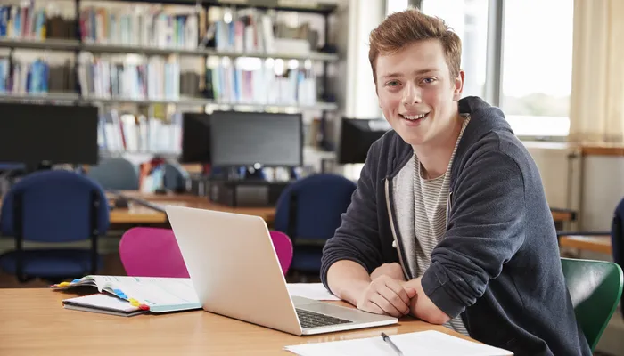 A young person sitting at a table in a library or study room, working on a laptop with open notebooks and papers nearby. Background includes bookshelves filled with books and several computer monitors on desks, indicating an academic environment