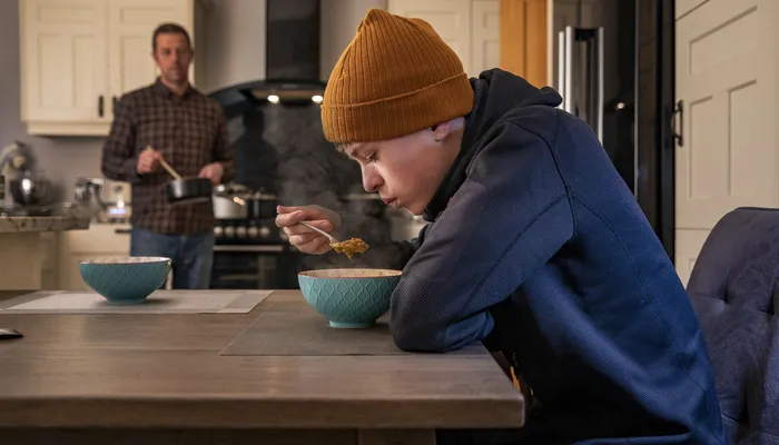 A kitchen scene showing a teenager wearing a mustard beanie and navy hoodie eating from a bowl at a wooden table, with an adult in a checked shirt cooking in the background. The setting highlights a casual, homey atmosphere with neutral-toned cupboards
