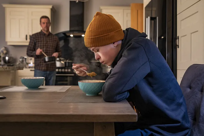 A kitchen scene showing a teenager wearing a mustard beanie and navy hoodie eating from a bowl at a wooden table, with an adult in a checked shirt cooking in the background. The setting highlights a casual, homey atmosphere with neutral-toned cupboards