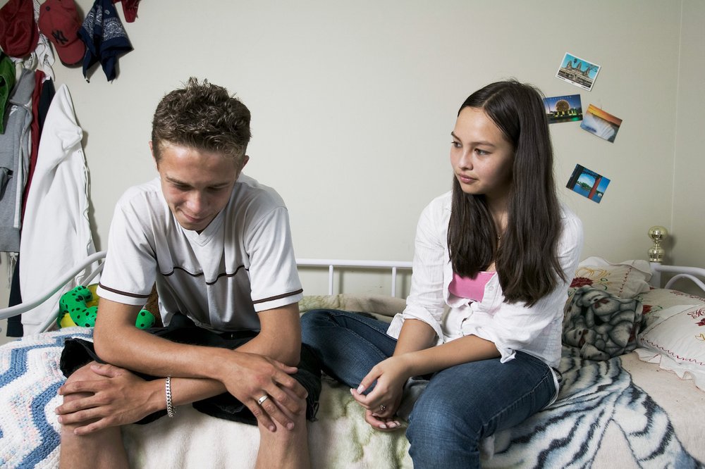 Teenage boy sitting on bed looking worried and talking to sister.