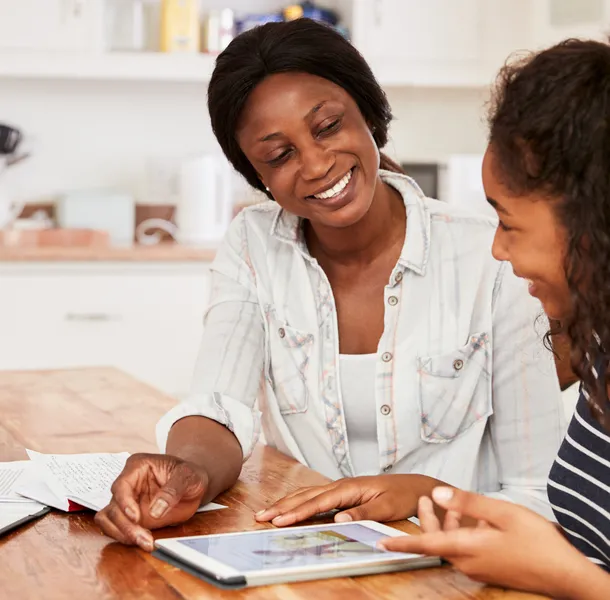 An adult and a teenager sitting at a wooden table in a kitchen, engaged in a discussion while looking at a tablet. The setting includes papers and a clipboard on the table, suggesting a collaborative or educational activity.