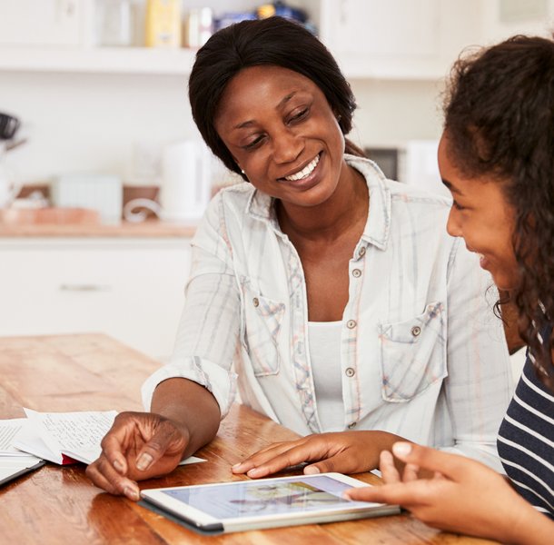 An adult and a teenager sitting at a wooden table in a kitchen, engaged in a discussion while looking at a tablet. The setting includes papers and a clipboard on the table, suggesting a collaborative or educational activity.