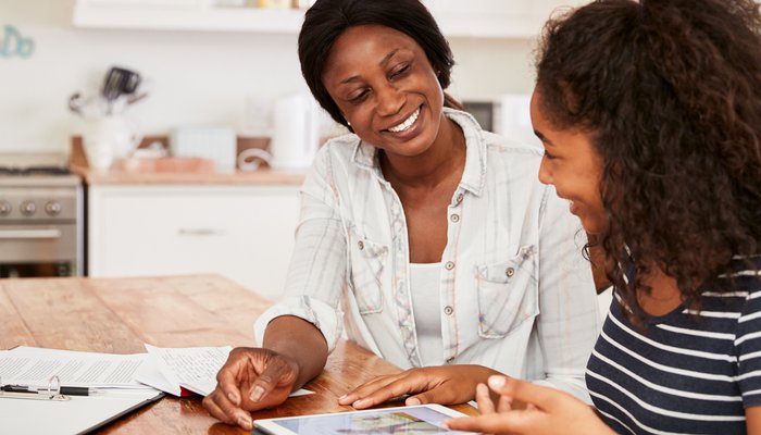 An adult and a teenager sitting at a wooden table in a kitchen, engaged in a discussion while looking at a tablet. The setting includes papers and a clipboard on the table, suggesting a collaborative or educational activity.
