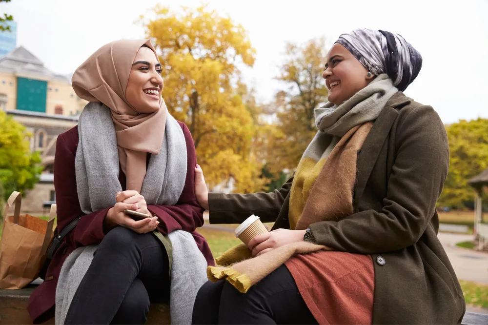 Two women wearing headscarves sitting on a bench outdoors during autumn, engaged in conversation. One woman holds a coffee cup while the other rests her hand on her companion's arm, with autumn foliage and a building visible in the background.