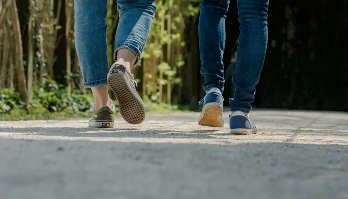 Lower legs and feet of two people walking on a gravel path surrounded by trees. One person wears blue jeans and green shoes, while the other wears darker blue jeans and blue shoes.