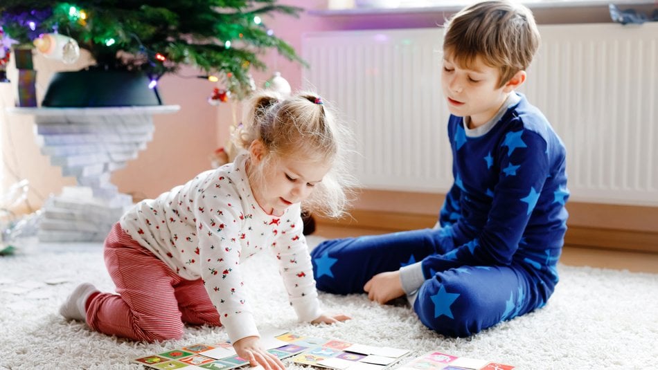 Two siblings, a little girl and boy, children sit on the floor at home playing together next to a decorated Christmas tree