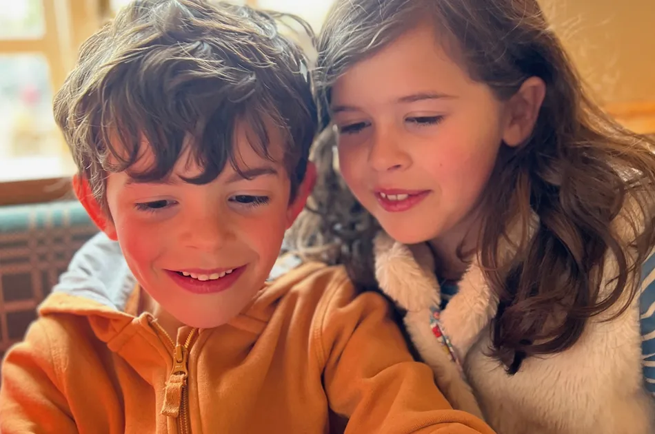Two children sitting closely together indoors, with one wearing an orange zip-up jacket and the other in a white furry gilet over a blue shirt. The setting appears casual and warm. They are looking at a tablet or device partially visible in the foreground